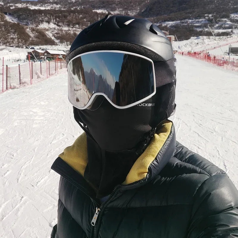 Rider on a ski slope wearing the black balaclava under a black helmet and goggles, with the neck tucked into a puffer jacket collar