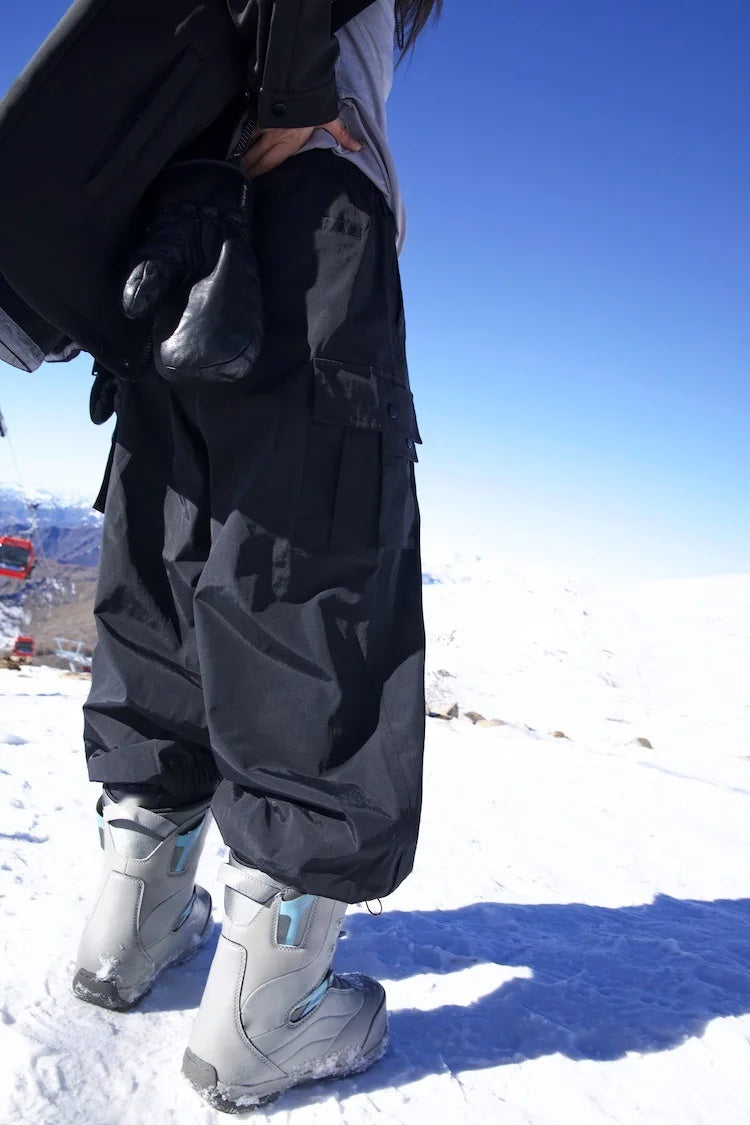 Sidehit Baggy Cargo Snow Pant in Black, worn on a snowy slope, side view of the thigh cargo pocket and gathered cuffs